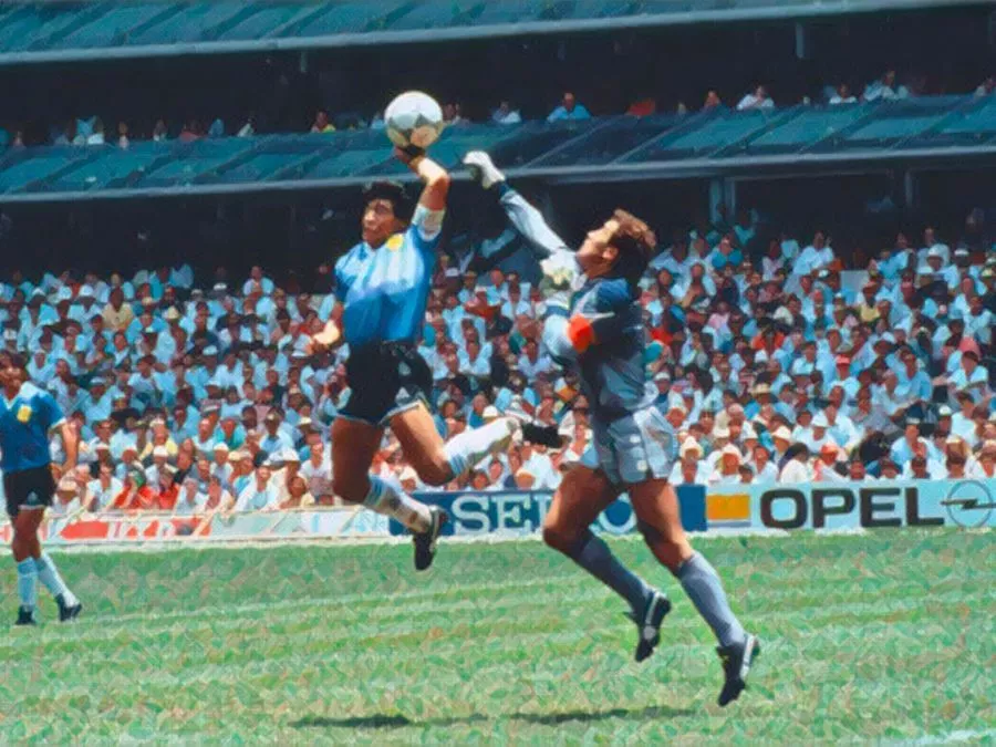 Diego Armando Maradona in maglia azzurra salta superando il portiere inglese Peter Shilton per colpire il pallone con la mano, segnando il gol della Mano de Dios all'Estadio Azteca nel 1986.